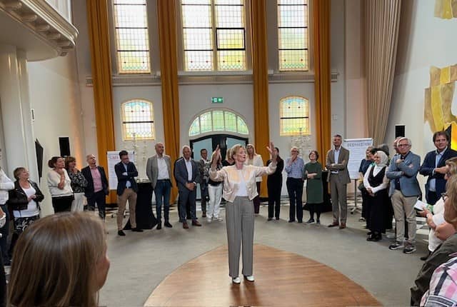 In mooie oude zaal heft vrouw haar armen omhoog staand in een kring van toehoorders