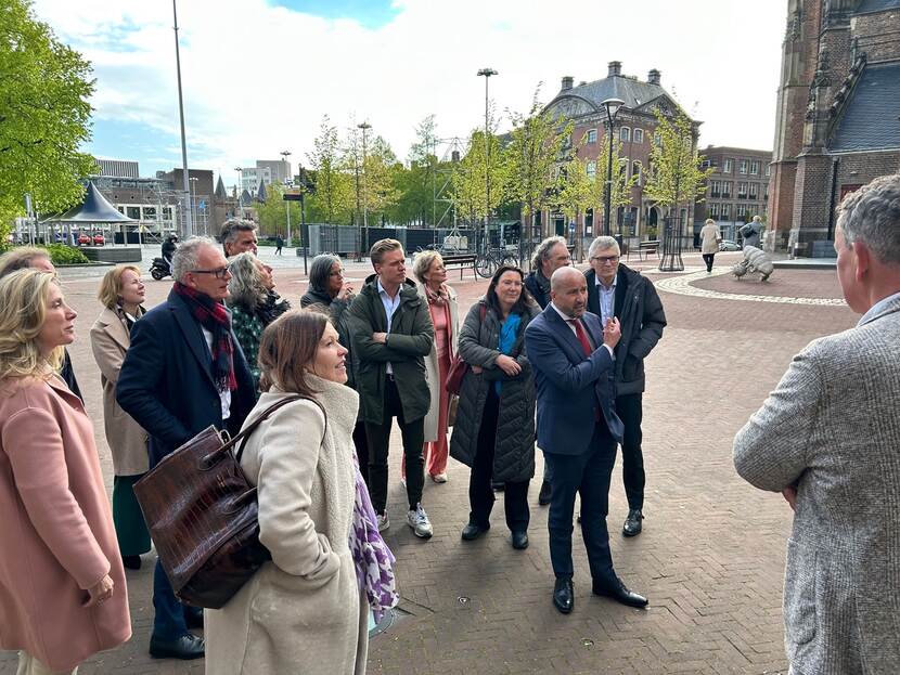 Zo'n 15 mensen staan buiten in een stad met een jas aan