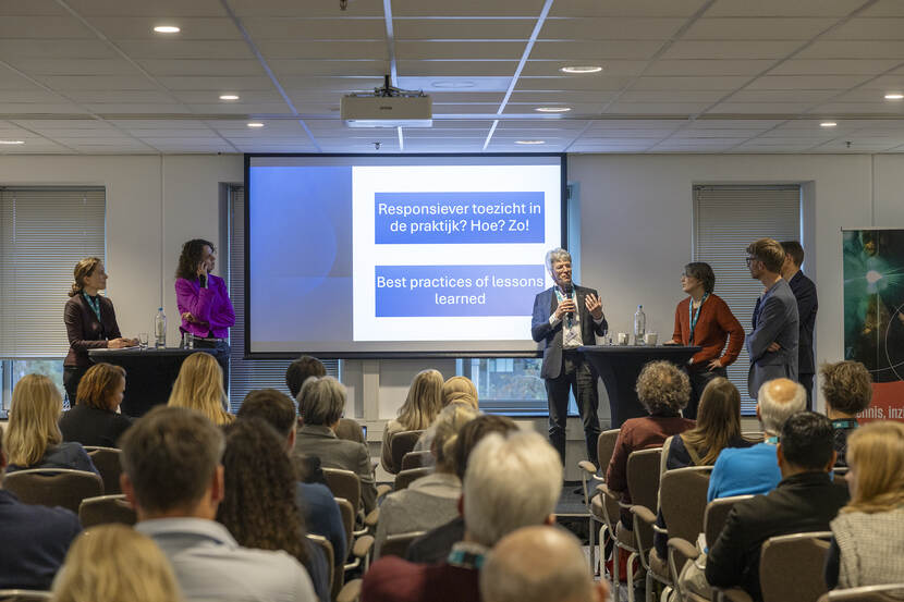 6 mensen staan voor een zaal met bezoekers van wie je alleen het achterhoofd ziet