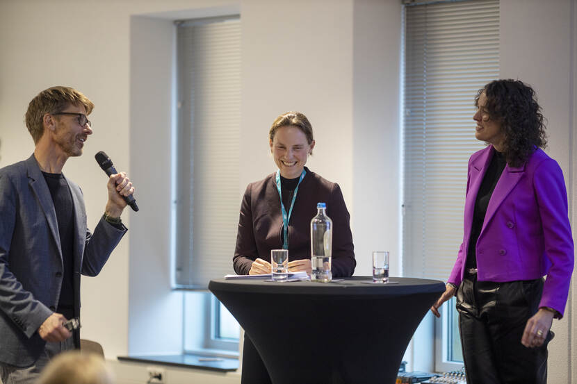 1 man met microfoon staat aan een tafel met 2 vrouwen