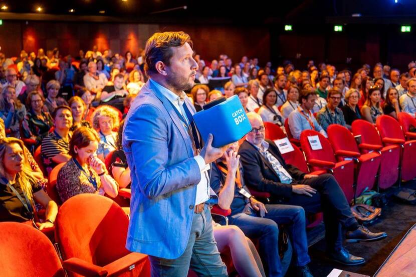 Man met blauw jasje heeft microfoon en stelt in de zaal een vraag