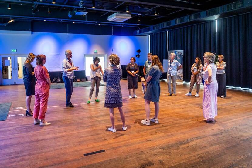 Op een houten vloer staan deelnemers aan een workshop in een grote kring om elkaar