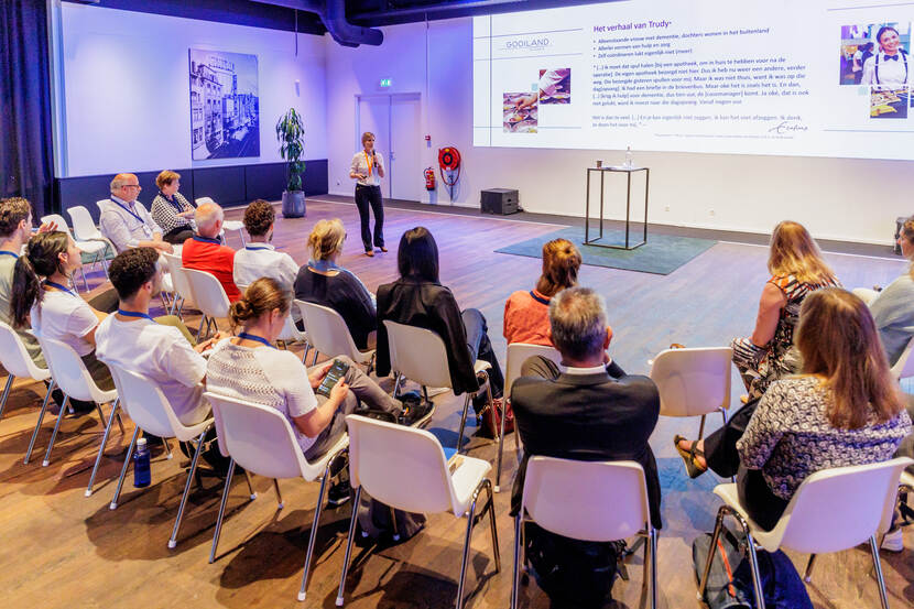 In een ruime zaal zitten 2 rijen met toehoorders te luisteren naar een man die een presentatie geeft