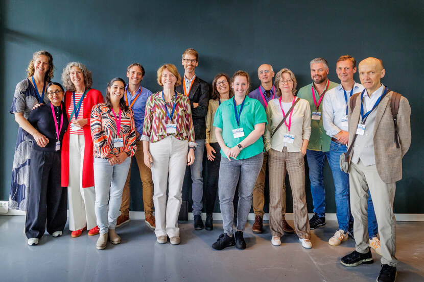 Een groep van 15 mensen kijkt staand naast elkaar de fotograaf aan
