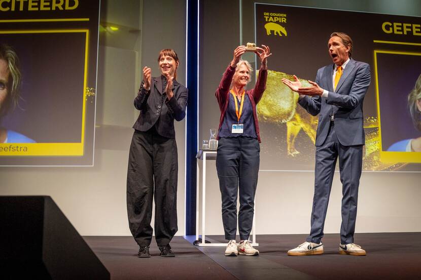 Op podium houdt een vrouw een beeldje omhoog, geflankeerd door andere vrouw en man