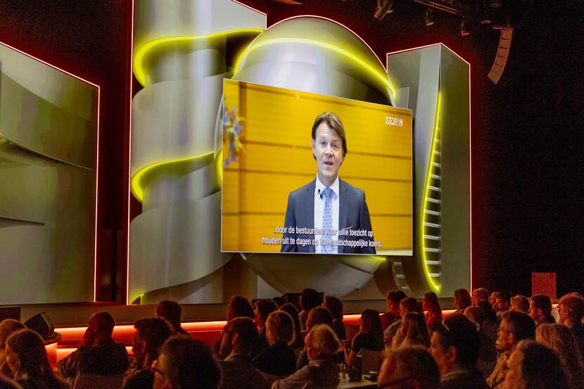 Mensen in een zaal kijken naar een filmpje waarin een man vertelt