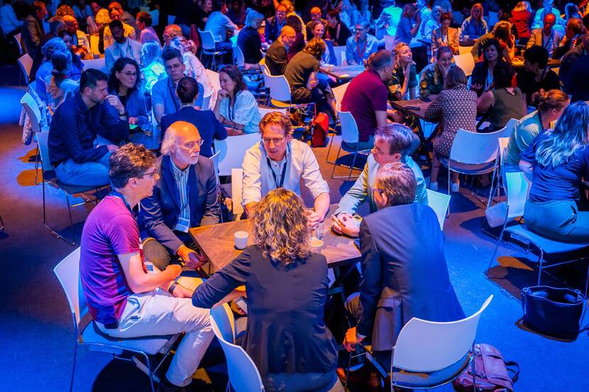In een blauw licht discussiëren 6 mensen aan een tafel te midden van een grotere zaal met andere mensen