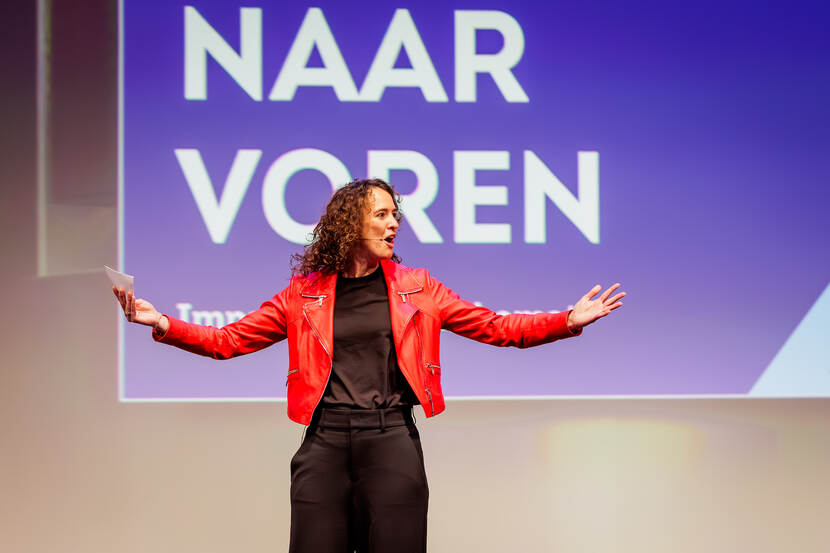 Vrouw in rood jasje met gespreide armen staat voor de tekst naar voren