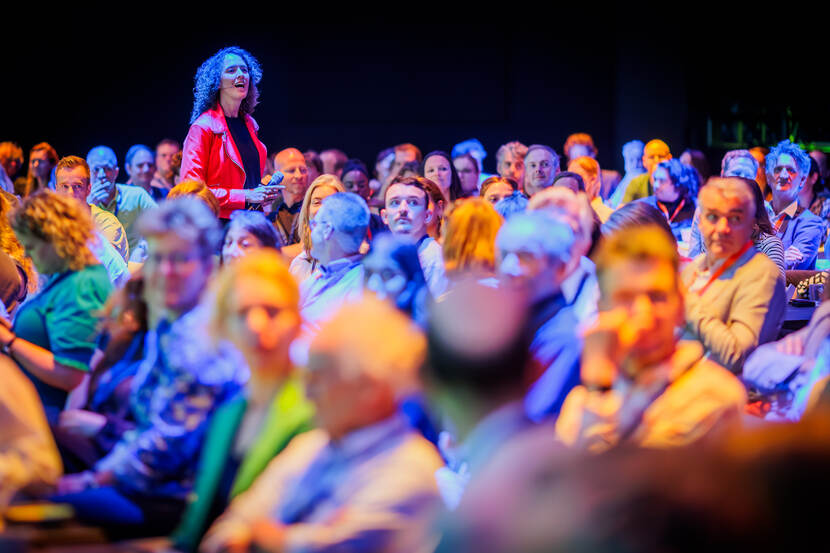 Vrouw in rood jasje steekt uit boven zaal met bezoekers